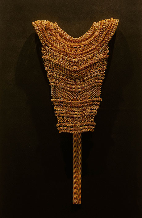 An intricate, golden chainmail-like garment hangs against a dark background, showcasing detailed loops and flowing texture. An intricate, golden chainmail-like garment hangs against a dark background, showcasing detailed loops and flowing texture.