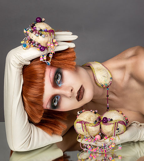 A model with vibrant red hair poses elegantly, adorned with unique, colorful jewelry pieces fashioned from baseballs and stylish white gloves. A model with vibrant red hair poses elegantly, adorned with unique, colorful jewelry pieces fashioned from baseballs and stylish white gloves.