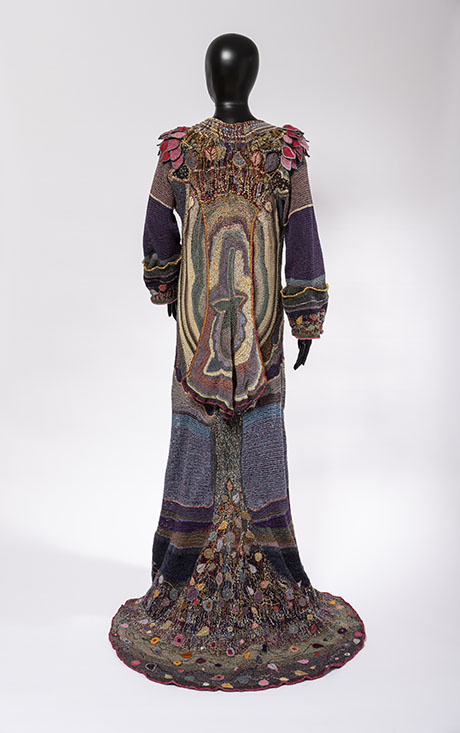 A colorful, intricately designed gown displayed on a black mannequin, featuring unique patterns and floral embellishments on the sleeves and hem. A colorful, intricately designed gown displayed on a black mannequin, featuring unique patterns and floral embellishments on the sleeves and hem.