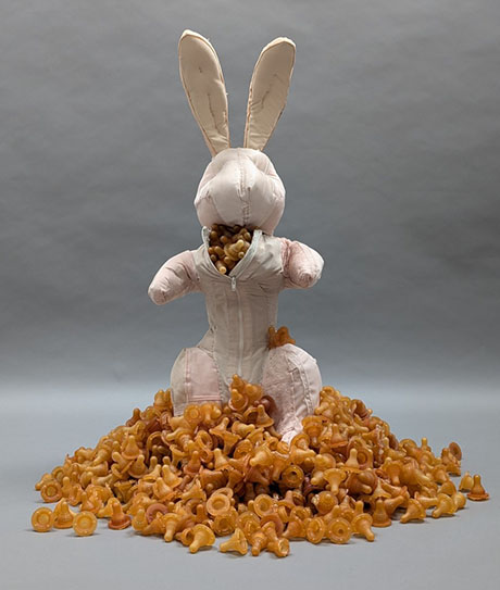 A surreal bunny figure wearing a pink jumpsuit sits amidst a mound of orange pasta shapes, exuding an odd, whimsical charm. A surreal bunny figure wearing a pink jumpsuit sits amidst a mound of orange pasta shapes, exuding an odd, whimsical charm.