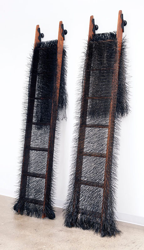 Two tall, wooden ladders draped with black, spiky, fur-like material, leaning against a white wall on a polished concrete floor. Two tall, wooden ladders draped with black, spiky, fur-like material, leaning against a white wall on a polished concrete floor.