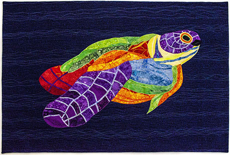 A colorful fabric art piece of a fish, featuring vibrant patterns of purple, green, yellow, and red, against a dark blue background.