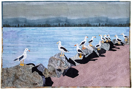 A quilt depicting seagulls perched on rocks by a calm water body, with mountains in the background and a sandy shoreline.