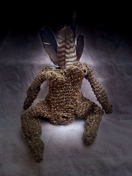 A handmade, brown crocheted figure with feathered accents sits against a softly lit background, evoking a mystical presence.