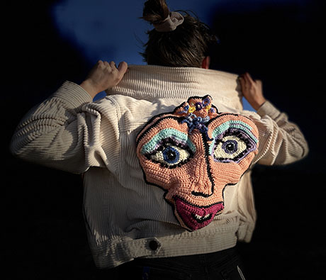 A person wearing a light-colored, oversized jacket, adjusting it on their shoulders against a dim, atmospheric backdrop.