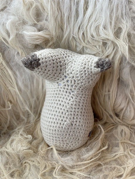 A handmade crocheted feminine form in light gray with dark gray tips, sitting on a soft, fluffy cream background.