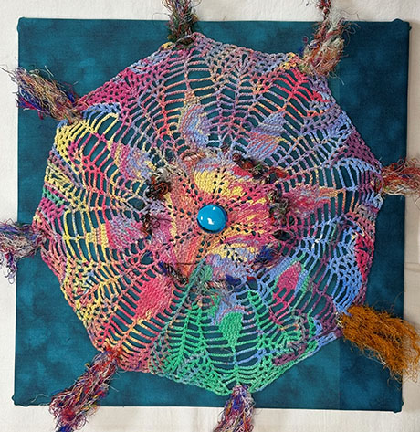 A vibrant, multicolored crochet mandala with a blue center button, adorned with fringes, is placed against a dark teal fabric, creating an artistic and lively tone.