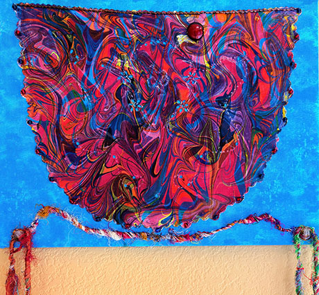 An abstract artwork with a vibrant, swirling mix of red, blue, and yellow marbling. The background is a bright blue with fringed, colorful yarn accents.