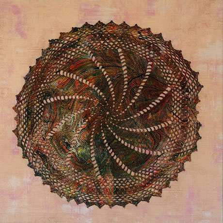 A circular, multicolored crochet doily with intricate patterns on a soft pink background. The design radiates a spiral effect, conveying a warm and artistic tone.
