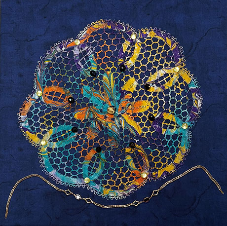 A vibrant, intricate circular textile art piece with colorful threads and beads on a deep blue background. A gold chain drapes underneath.