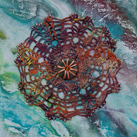 A vibrant, circular crochet pattern in earthy hues overlays a swirling teal and white marbled background. A central, textured button completes the design.