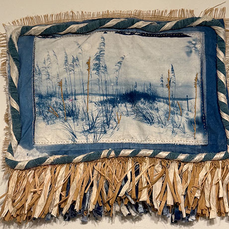 Textile art piece featuring a blue-toned landscape of tall grass on a snowy field, bordered by a blue frame and fringed with woven fabric strips.