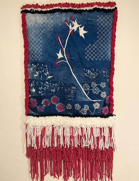Textile art piece featuring a floral design with white flowers and red accents on a blue background. Red and white fringe decorates the bottom.