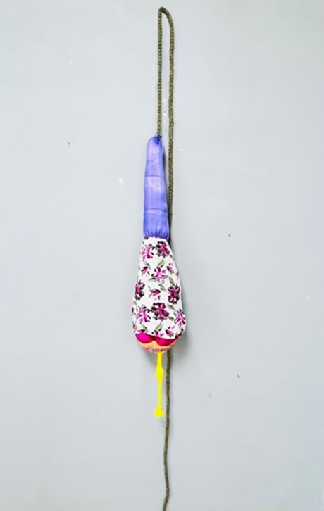 A colorful hanging bag against a gray background. The top is purple, and the bottom is white with pink floral patterns. A yellow tassel hangs below.