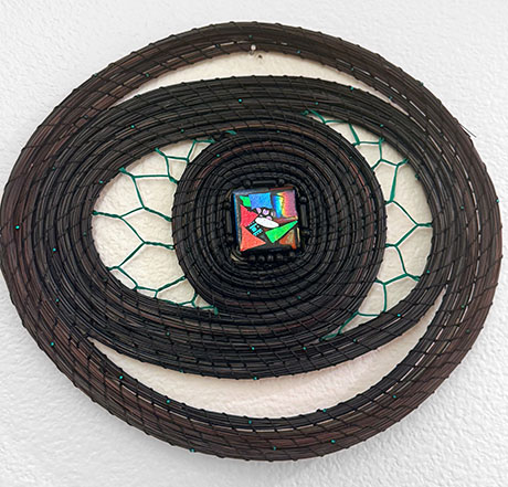 A spiraled sculpture with a colorful glass tile at the center, set against a textured white wall. The piece conveys a modern, intricate, and vibrant feel.