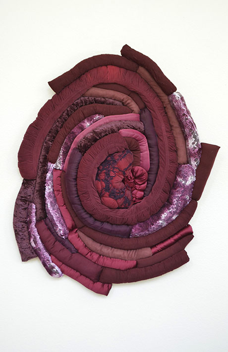 Textile art resembling a rose, with layered, textured fabrics in rich red, burgundy, and pink hues. The intricate swirls create a sense of depth and warmth.