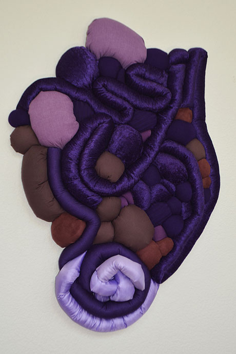 Abstract textile art with layered purple, pink, and brown plush shapes, resembling organic forms. The composition is soft and visually tactile.