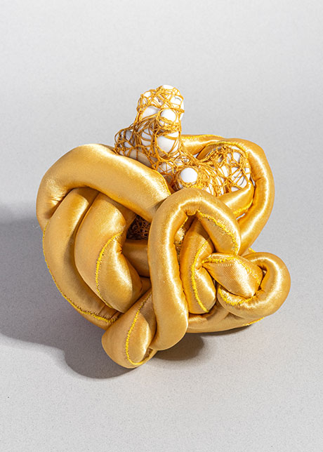 A complex yellow sculpture features knotted, satin-covered tubes intertwined with a delicate, web-like mesh. The structure evokes curiosity and creativity.