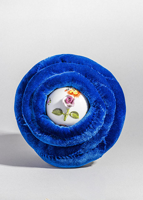 A porcelain ball with floral patterns is encased in vibrant blue velvet folds, resembling a blooming flower. The tone is elegant and luxurious.