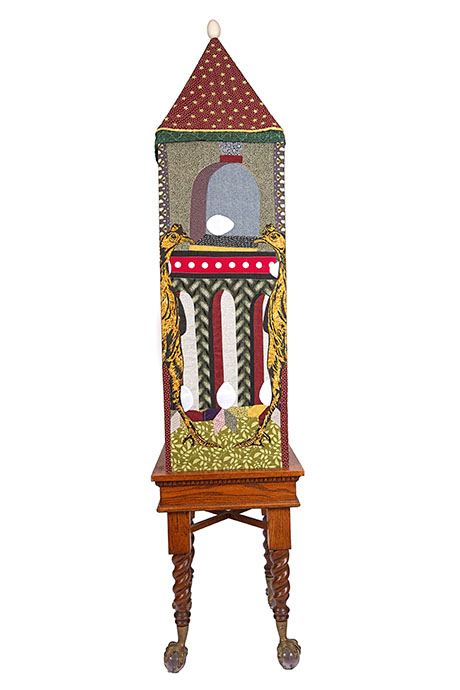 An intricately designed sculpture with a colorful, fabric-covered obelisk on a wooden base. The obelisk features vibrant patterns and chicken-and-egg motifs.