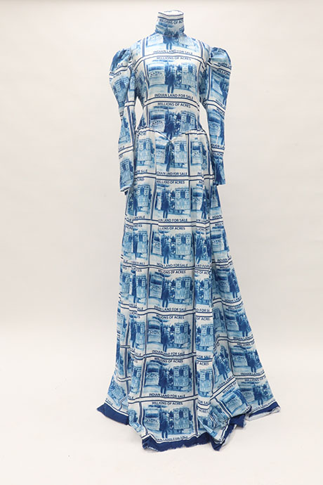 Blue and white floor-length dress with historical "Indian Land for Sale" print, featuring puffed sleeves and a high neck, against a plain backdrop.