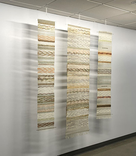 Three vertical woven tapestries with earthy tones hang on a white gallery wall. Each tapestry features intricate patterns, creating a serene and artistic ambiance.