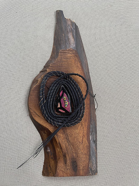 A colorful embroidered coil rests on a smooth, polished piece of wood against a textured gray background, creating a rustic and artistic contrast.