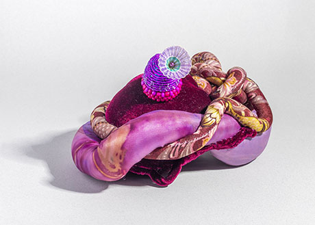 A vibrant sculptural piece featuring intertwined fabric twists in shades of pink and maroon, topped with a beaded purple spiral and a gem-like center.