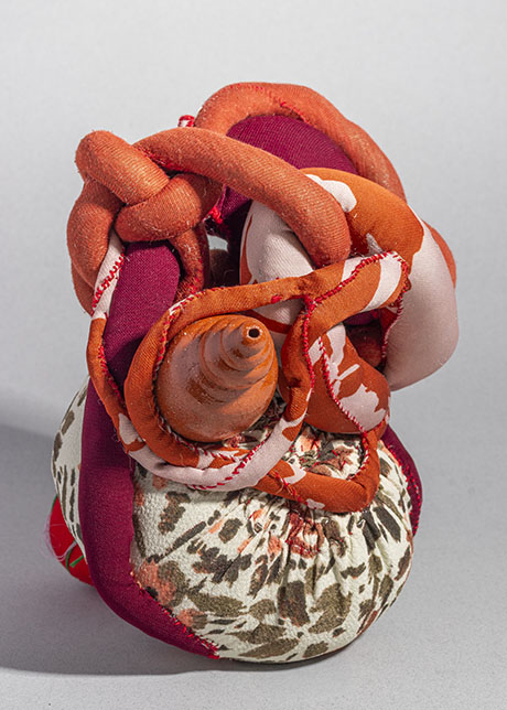 Abstract textile sculpture with intertwined swirling fabrics in orange, red, and pink hues atop a floral-patterned base, evoking a sense of dynamic motion.