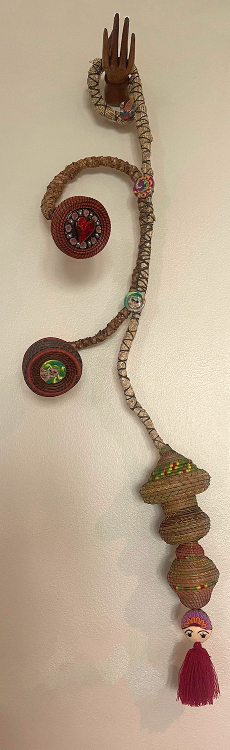 A decorative wall hanging featuring a curly, woven rope with attached wooden elements, colorful beads, and small figures. It ends with a pink tassel.