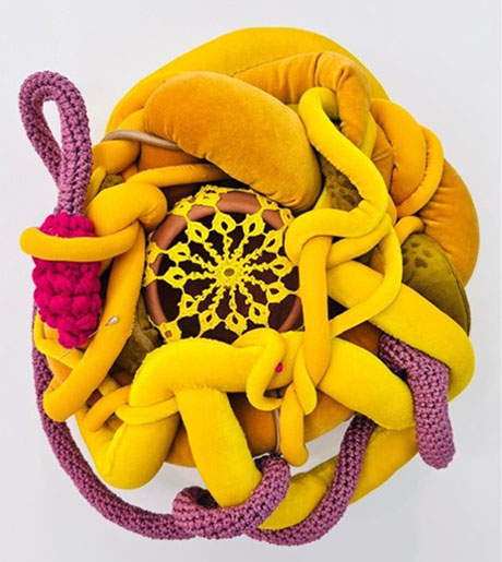 Colorful textile sculpture with intertwined ropes and tubes in yellow and pink, resembling a flower. Central crochet pattern adds intricate detail.