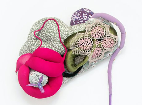 Abstract textile art piece with layered textures: pink cushions, crochet lace, green and gray patterns, and purple accents on a white backdrop.