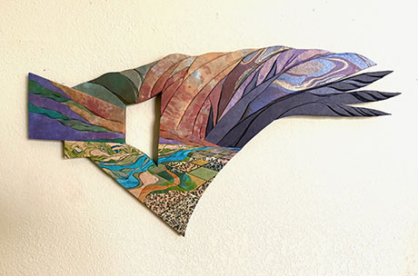 Abstract wall sculpture with layered, jagged shapes in earthy tones of green, brown, and purple. Suggests a dynamic landscape or mountain silhouette becoming a wing with feathers.