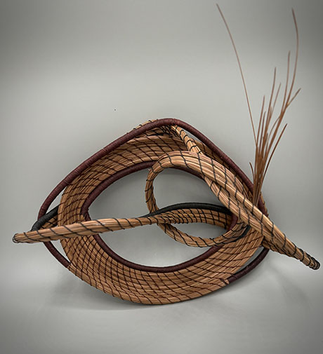 A modern abstract sculpture made of looped and intertwined natural fibers, creating a dynamic, flowing form on a neutral gray background.