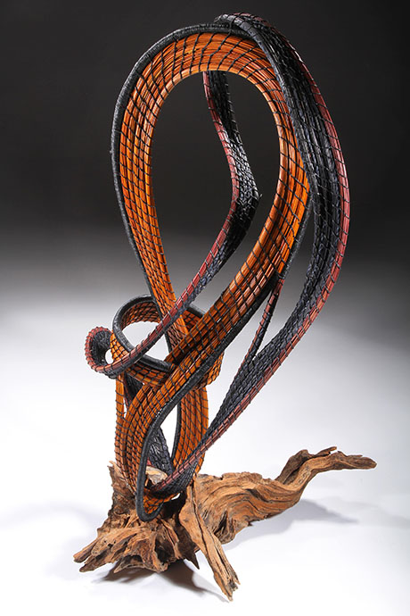Intricately looped sculpture with orange and black woven patterns spirals upward from a textured wooden base. The design is dynamic and abstract.