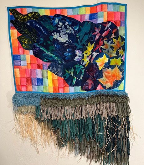 Textile artwork with vibrant leaves against a colorful patchwork background. Below, wool fringe cascades in earth tones, evoking a sense of nature's flow.