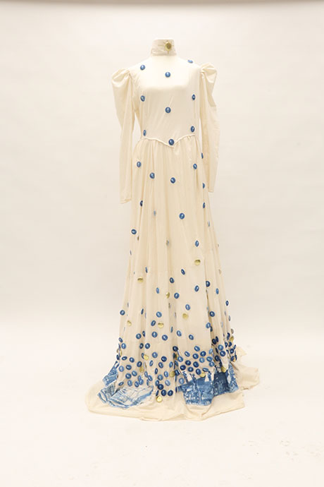 Elegant white gown with a high collar and long sleeves, adorned with scattered blue discs. The hem features a blue artistic pattern. Sophisticated and modern.