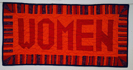 A vibrant quilted banner with "WOMEN" in bold red letters on an orange background, surrounded by a striped red and purple-black border, hangs on a white wall.