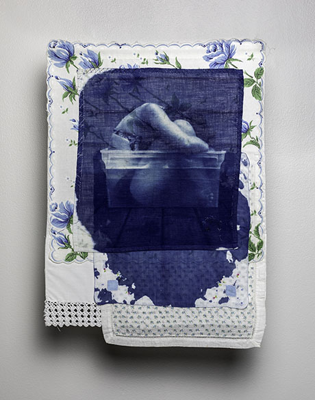 A blue-toned image of a person bending over a tub is printed on a floral cloth with lace trim. The artwork has a vintage and intimate feel.