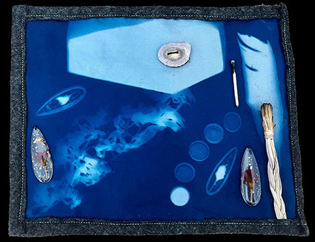 Blue textured art piece with abstract patterns, feathers, and organic shapes. A small geode, matchstick, and bundle of white twigs enhance its mystic feel.