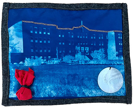 Textile artwork features a cyanotype image of a building on blue fabric, accented with a red fabric bow and a circular white patch, creating a vintage, artistic vibe.