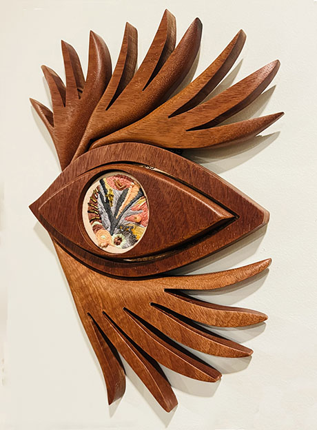 A wooden wall art piece resembling an eye with stylized, protruding brow and lashes, featuring an intricate embroidered inner design; language of spiritual exploration and generational dialogue