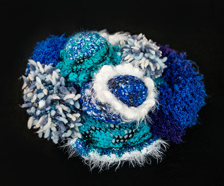 Close-up of a textured, multicolored blue and white knitted or crocheted abstract textile art piece by Marita Setas Ferro on a black background.