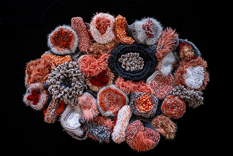 Abstract textile art composed of various textured, circular shapes in warm tones like orange, brown, and black on a black background.