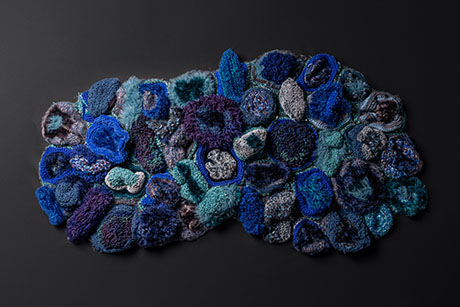 Abstract textile artwork composed of layered, textured blue and teal fabric pieces arranged on a dark background.