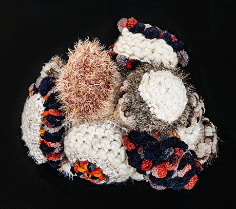 A vibrant, textured sculpture by Marita Setas Ferro of woven yarns in shades of white, beige, orange, and blue, resembling a cluster of coral. Organic and soft yet dynamic and intricate.