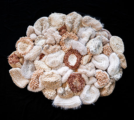 A textured circular art piece made of various crocheted and knitted shapes in beige, cream, and brown tones, set against a black background.