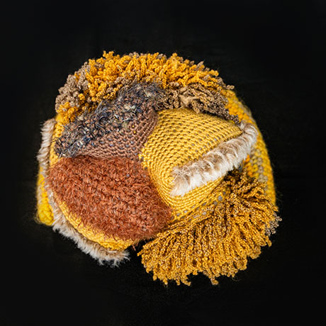 Multicolored textured textile art piece by Marita Setas Ferro featuring various knitted and looped yarn patterns in yellow, brown, and beige tones on a black background.