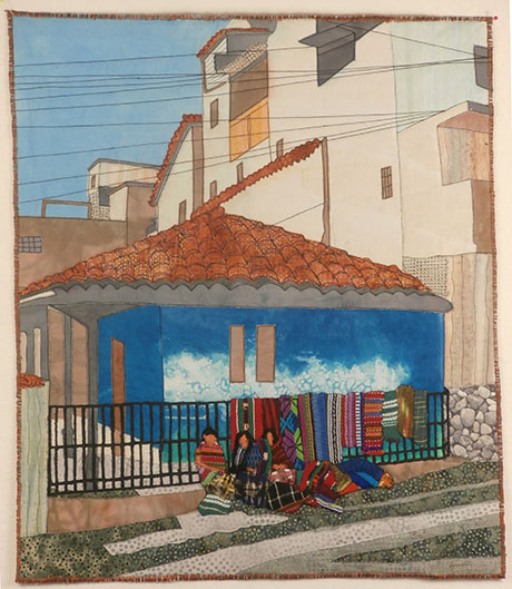 Quilt by Kathleen McCabe depicting a blue house with a red tile roof, women selling colorful textiles outside on a sloped street.