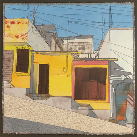 Quilted artwork by Kathleen McCabe depicting colorful hillside houses; bright yellow and orange walls under a blue sky. Textured cobblestone path adds depth.